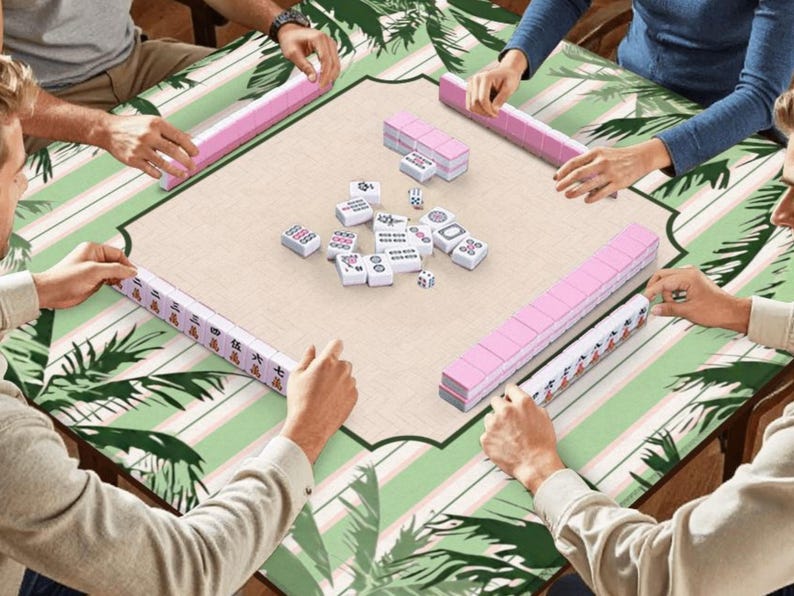 K&ouml;nnte beinhalten: Eine Mahjong-Partie, bei der Spieler um einen Tisch sitzen. Der Tisch hat ein gr&uuml;nes und wei&szlig;es tropisches Bl&auml;ttermuster. Rosa Kachelregale und Kacheln sind angeordnet, zusammen mit W&uuml;rfeln. Die Spieler halten die Kacheln.