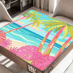 May include: A wooden table with a colorful tropical beach scene printed on the top. The design features palm trees, pink and green leaves, a sandy beach, and blue ocean waves. The table is surrounded by two chairs.