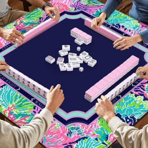 Puede incluir: Una vibrante partida de Mahjong en curso. La mesa presenta una superficie de juego azul marino con bordes florales rosas y turquesas. Se ven estanter&iacute;as rosas para fichas y fichas blancas con s&iacute;mbolos negros, junto con dados. Los jugadores interact&uacute;an con el juego.
