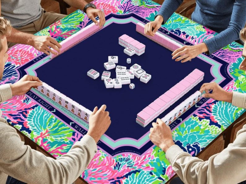 Puede incluir: Una vibrante partida de Mahjong en curso. La mesa presenta una superficie de juego azul marino con bordes florales rosas y turquesas. Se ven estanter&iacute;as rosas para fichas y fichas blancas con s&iacute;mbolos negros, junto con dados. Los jugadores interact&uacute;an con el juego.