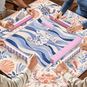 Pu&ograve; includere: Un tavolo da gioco Mahjong quadrato con un design a onde blu e bianche, circondato da tessere rosa e bianche. Il tavolo &egrave; posto su un tavolo con un motivo floreale e di conchiglie. Diverse persone stanno giocando.