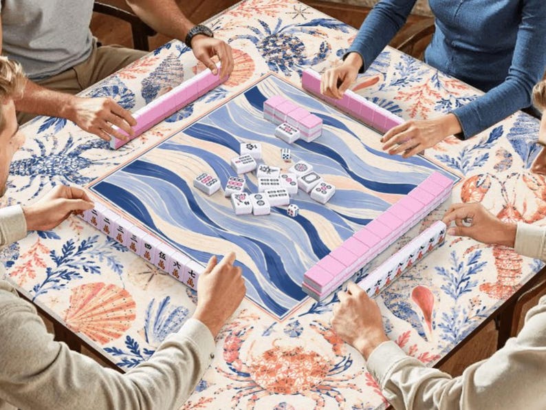 Pu&ograve; includere: Un tavolo da gioco Mahjong quadrato con un design a onde blu e bianche, circondato da tessere rosa e bianche. Il tavolo &egrave; posto su un tavolo con un motivo floreale e di conchiglie. Diverse persone stanno giocando.