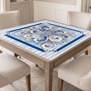 May include: A square wooden table with a watercolor oyster design on the tabletop. The design features blue, purple, and white oyster shells arranged in a circular pattern on a blue background with blue and white stripes. Four beige chairs surround the table.