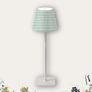 May include: A white table lamp with a mint green patterned lampshade. The lamp has a tall, slender base and a square base. Several Mahjong tiles are scattered around the base of the lamp.