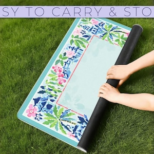 May include: A rolled-up yoga mat with a floral and palm tree design is held by two hands on a grassy surface. The mat has a light blue background with pink and blue floral accents. The text "EASY TO CARRY & STORE" is at the top.