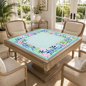 Pu&ograve; includere: Un tavolo quadrato in legno con una superficie azzurra e un bordo floreale rosa, blu e verde. Il tavolo &egrave; circondato da quattro sedie in vimini con cuscini beige. La stanza ha grandi finestre e un tema tropicale.