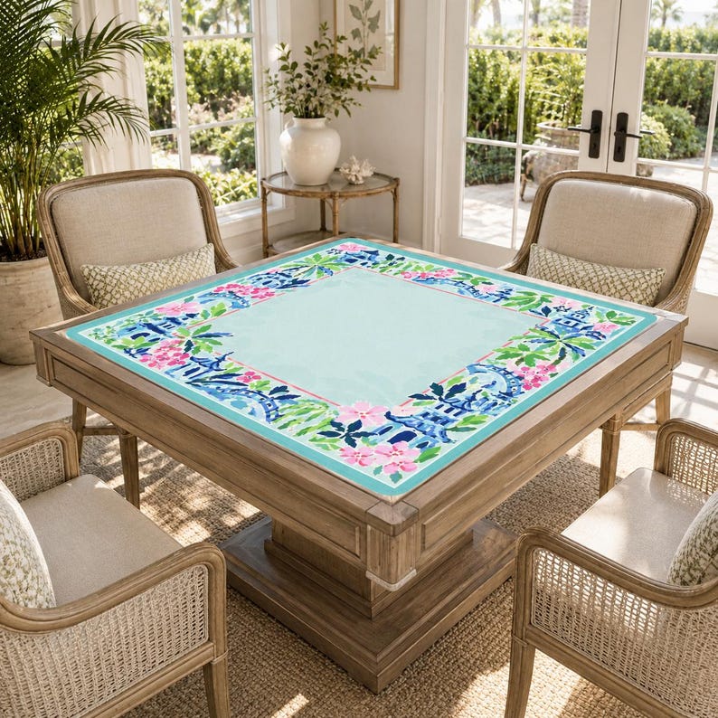 Pu&ograve; includere: Un tavolo quadrato in legno con una superficie azzurra e un bordo floreale rosa, blu e verde. Il tavolo &egrave; circondato da quattro sedie in vimini con cuscini beige. La stanza ha grandi finestre e un tema tropicale.