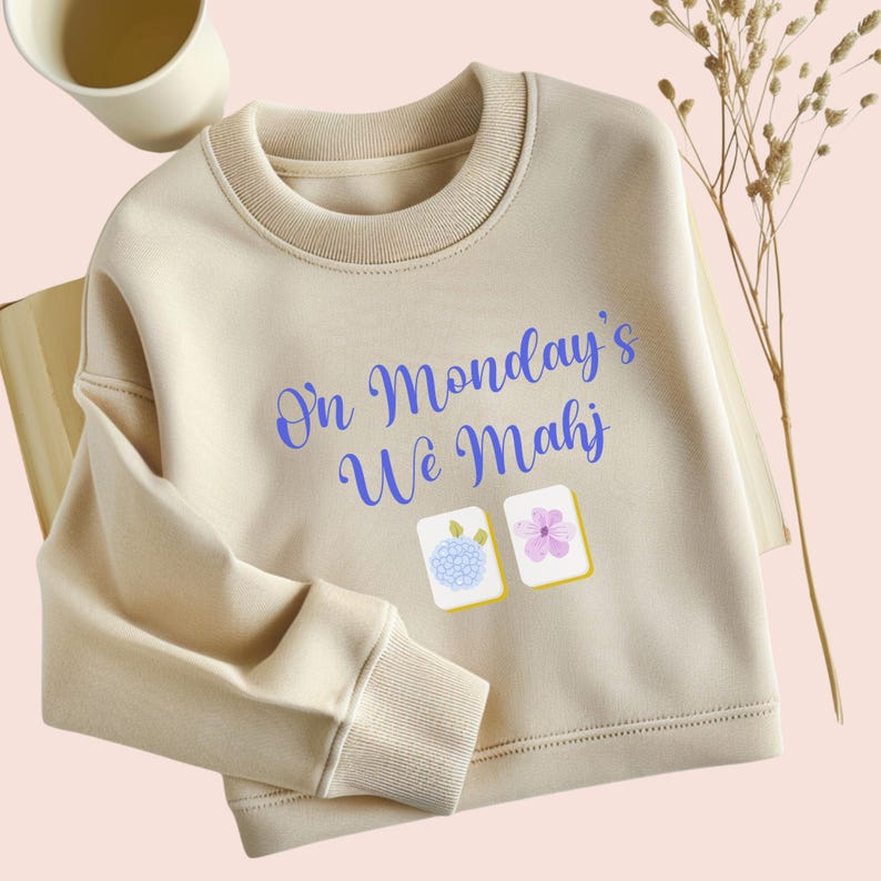 May include: Cream-colored sweatshirt with the text "On Monday's We Mahj" in blue script. Two small images of Mahjong tiles are below the text. The sweatshirt has a ribbed crew neck and cuffs.