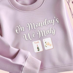 May include: A light purple sweatshirt with the text "On Monday's We Mahj" in white script. Two small square graphics show a champagne bottle and a flower. The sweatshirt has a crew neck and ribbed cuffs.