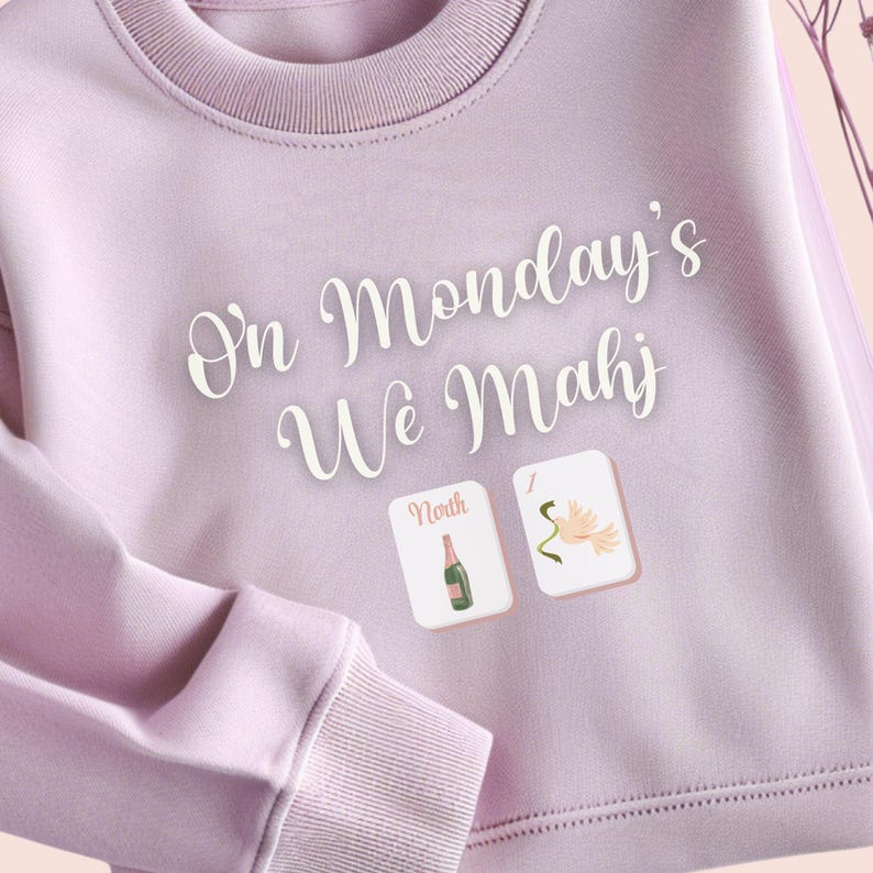 May include: A light purple sweatshirt with the text "On Monday's We Mahj" in white script. Two small square graphics show a champagne bottle and a flower. The sweatshirt has a crew neck and ribbed cuffs.