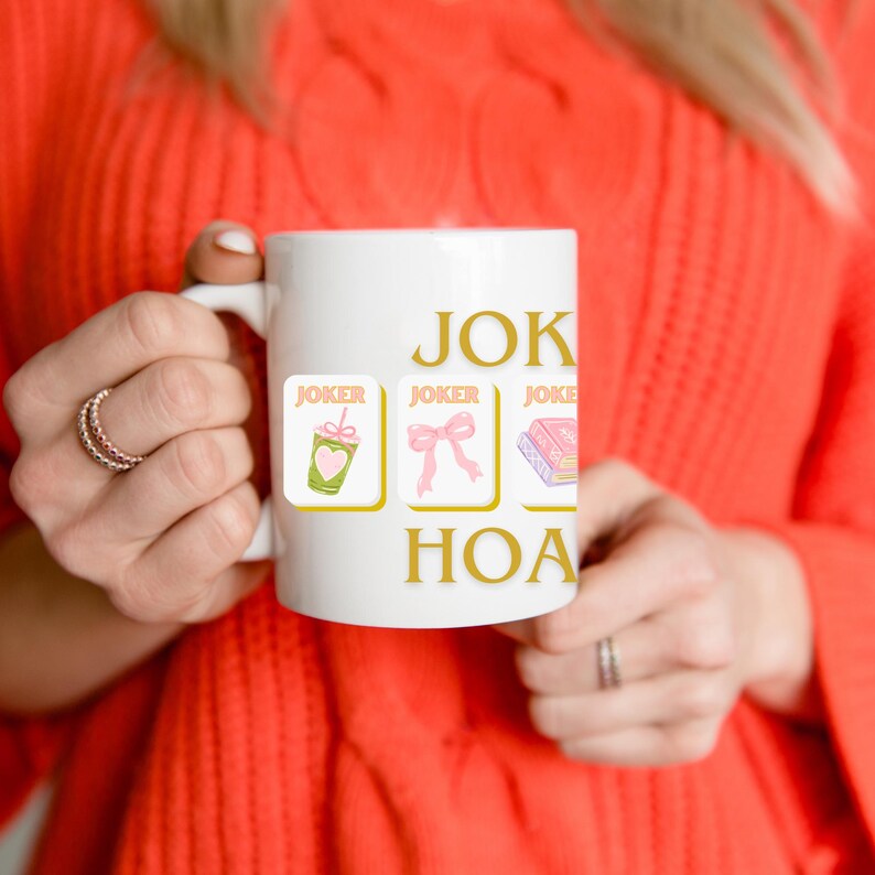 May include: White ceramic mug with the words "JOKER HOA" in gold lettering. The mug features three small square images with the word "JOKER" above each. The images include a green drink, a pink bow, and a pink book.