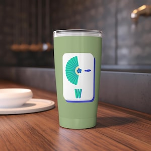 May include: A sage green tumbler with a white rectangle featuring a teal fan design and the letter "W". The fan has a blue flower and a blue line. The tumbler is on a wooden surface.