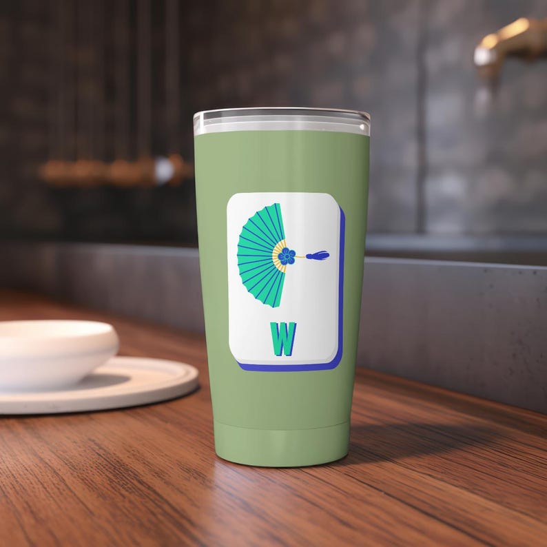 May include: A sage green tumbler with a white rectangle featuring a teal fan design and the letter "W". The fan has a blue flower and a blue line. The tumbler is on a wooden surface.