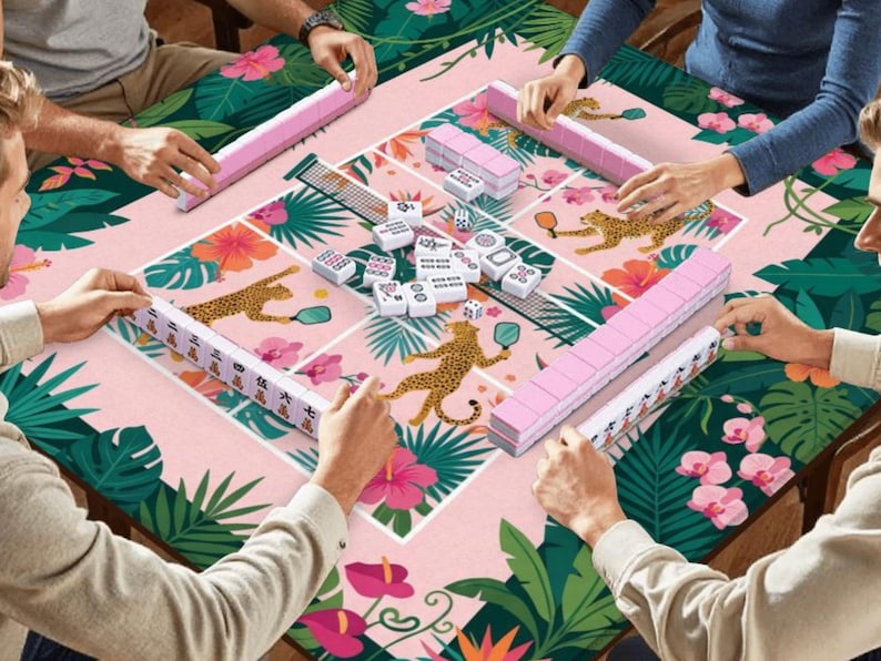 May include: A group of people playing Mahjong on a table with a pink floral and leopard print design. The game pieces are white and pink. The table is surrounded by green tropical leaves and pink flowers.
