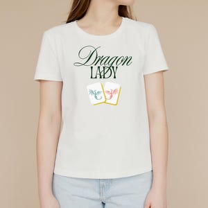 May include: White t-shirt with the text "Dragon Lady" in green and black script. Below the text are two small cards with dragon illustrations in blue and red. The shirt is a crew neck style.