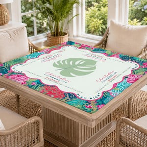 May include: A square table with a decorative top featuring a tropical floral design in pink, teal, and green. The table has a light wood frame and is surrounded by wicker chairs. The table top has the word "Charleston" and a large green leaf.