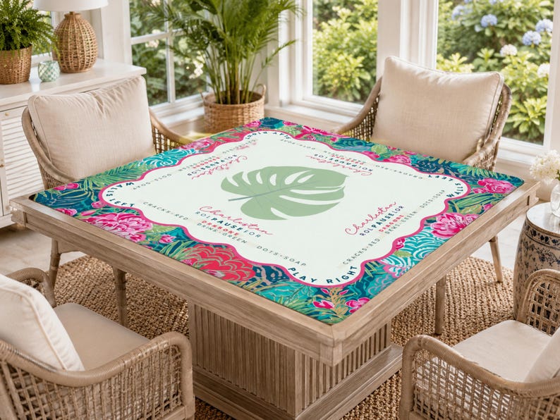 May include: A square table with a decorative top featuring a tropical floral design in pink, teal, and green. The table has a light wood frame and is surrounded by wicker chairs. The table top has the word "Charleston" and a large green leaf.