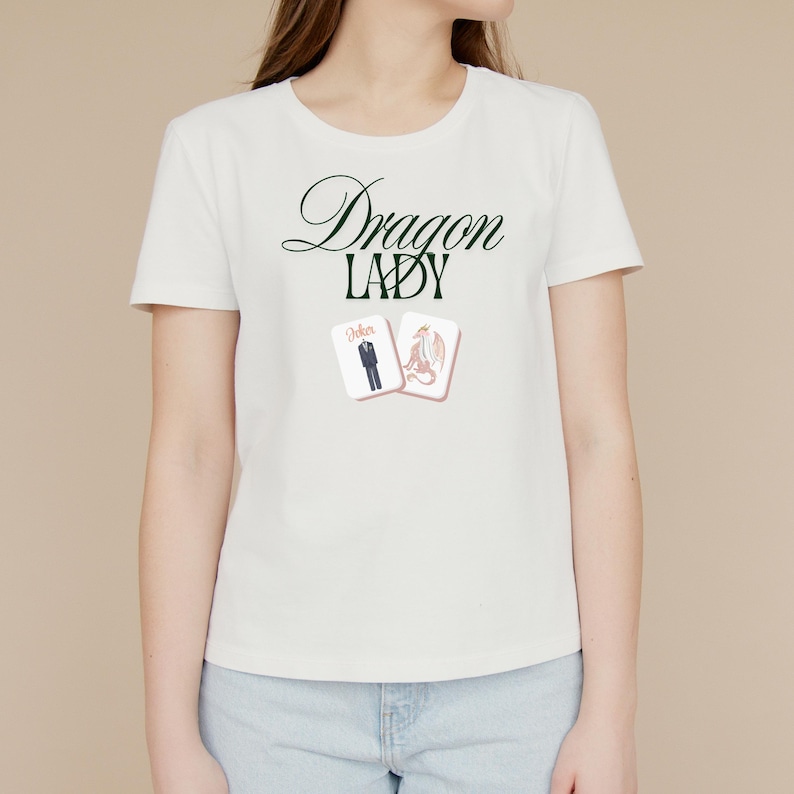 May include: White t-shirt with the text "Dragon Lady" in a dark green script. Below the text are two playing cards, one with a suit and the other with a dragon illustration. The shirt is a crew neck style.