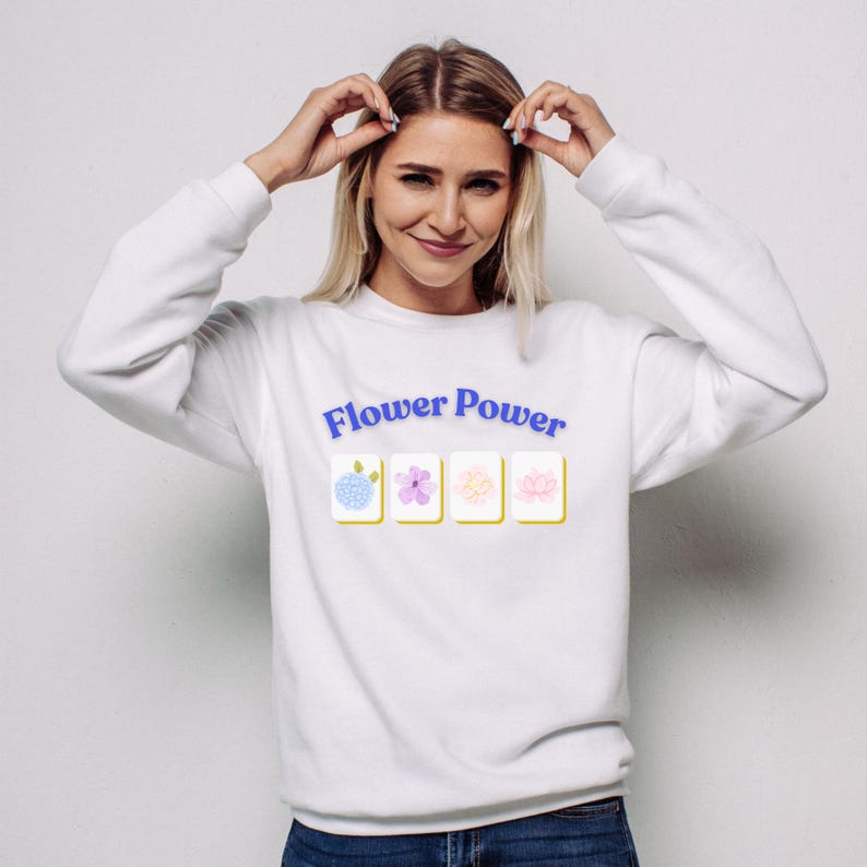 May include: White sweatshirt with the text "Flower Power" in blue, above four flower illustrations. The illustrations are in pastel colors, including blue, pink, and yellow. The sweatshirt is a casual style.