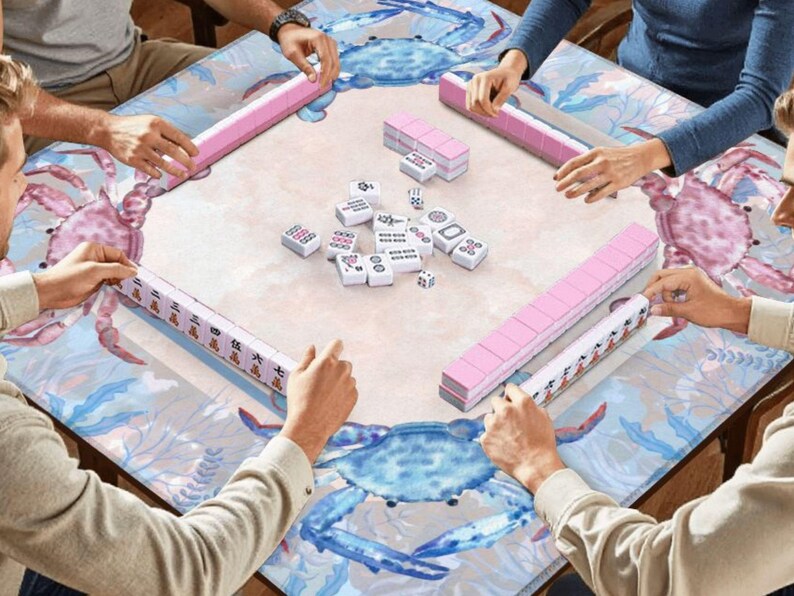 Puede incluir: Una mesa de Mahjong cuadrada con un dise&ntilde;o de cangrejo y oc&eacute;ano. Los jugadores est&aacute;n sentados alrededor, sosteniendo estantes de fichas rosas. Las fichas y dados de Mahjong blancos est&aacute;n en el centro, listos para jugar.