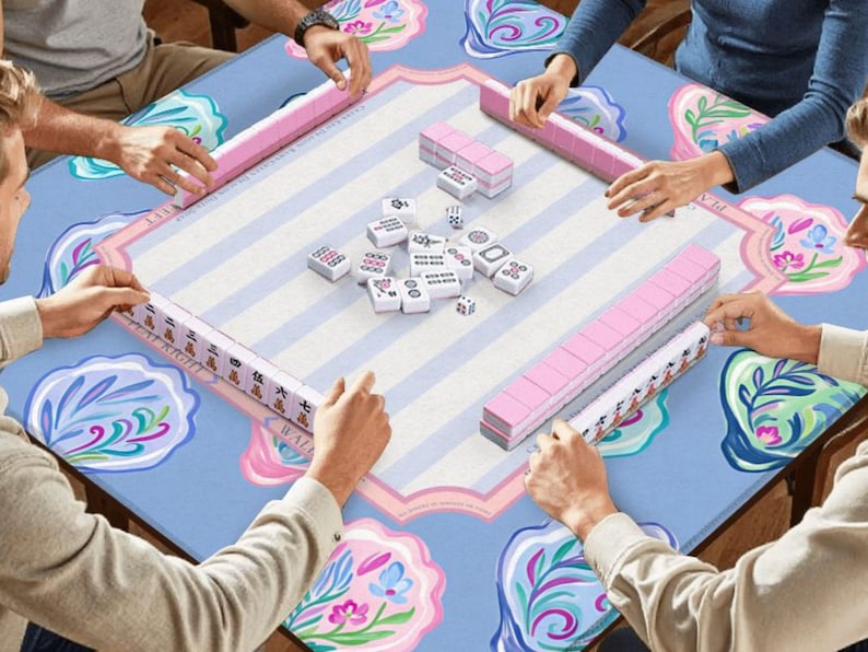 Puede incluir: Una partida de Mahjong en curso, con jugadores alrededor de una mesa. La mesa tiene un patr&oacute;n floral azul claro. Se ven estanter&iacute;as rosas y fichas blancas. Los jugadores sostienen las fichas y las estanter&iacute;as.