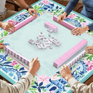 Pu&ograve; includere: Un gruppo di persone gioca a Mahjong. Il gioco &egrave; ambientato su un tavolo azzurro con un design floreale e blu. Sono visibili rastrelliere rosa per tessere e tessere bianche con segni neri.