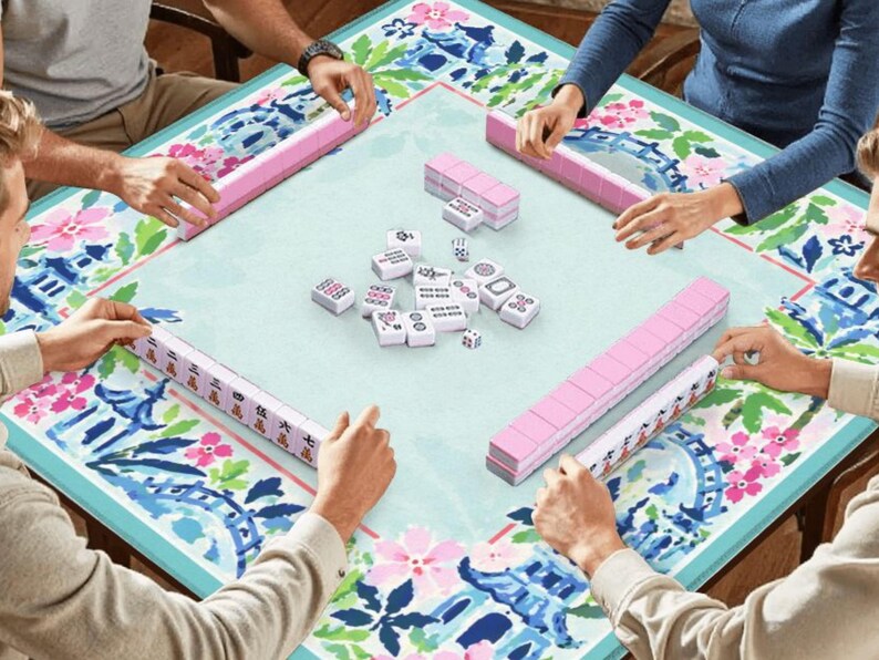 Pu&ograve; includere: Un gruppo di persone gioca a Mahjong. Il gioco &egrave; ambientato su un tavolo azzurro con un design floreale e blu. Sono visibili rastrelliere rosa per tessere e tessere bianche con segni neri.