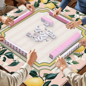May include: A group of people playing Mahjong on a table with a floral and lemon pattern. The game features pink tile racks and white tiles with black symbols. The table has a light pink background with yellow lemons and white flowers.
