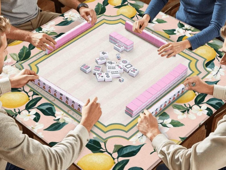 May include: A group of people playing Mahjong on a table with a floral and lemon pattern. The game features pink tile racks and white tiles with black symbols. The table has a light pink background with yellow lemons and white flowers.