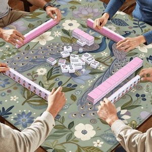 May include: A group of people playing Mahjong on a floral patterned table. The game features pink tiles and racks, with white tiles and dice scattered on the table. The table has a peacock and floral design.