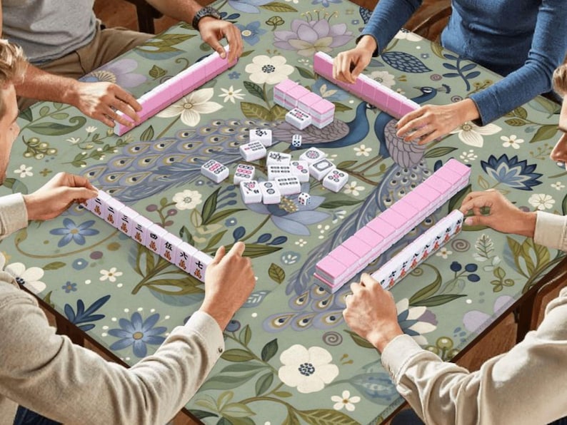 May include: A group of people playing Mahjong on a floral patterned table. The game features pink tiles and racks, with white tiles and dice scattered on the table. The table has a peacock and floral design.