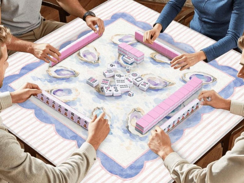 May include: A group of people playing Mahjong. The game is set on a square table with a light blue and white patterned surface. Pink tiles and racks are visible, along with other game pieces.