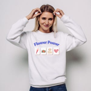 May include: White sweatshirt with the text "Flower Power" in blue. Below the text are four small images: a pink umbrella, a pumpkin, a stack of pancakes, and a heart made of roses. The model is wearing blue jeans.