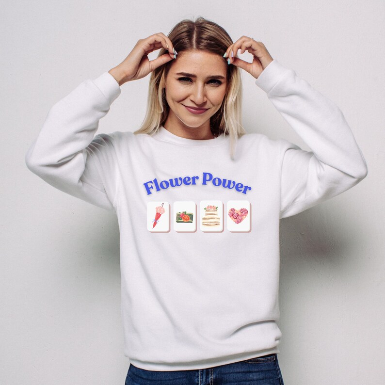 May include: White sweatshirt with the text "Flower Power" in blue. Below the text are four small images: a pink umbrella, a pumpkin, a stack of pancakes, and a heart made of roses. The model is wearing blue jeans.