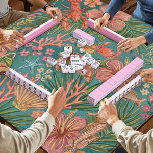 May include: A group of people playing Mahjong. The teal tablecloth has a floral and marine life design. Pink tile racks and white tiles are on the table. The game is in progress.