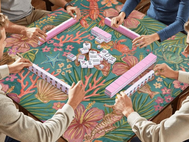 May include: A group of people playing Mahjong. The teal tablecloth has a floral and marine life design. Pink tile racks and white tiles are on the table. The game is in progress.