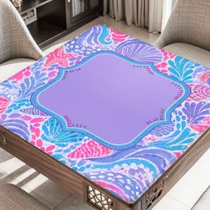K&ouml;nnte beinhalten: Ein quadratischer Tisch mit einer bunten, gemusterten Tischplatte. Das Design zeigt eine lila Mitte mit einem dekorativen Rand, umgeben von einer lebendigen Mischung aus rosa, blauen und wei&szlig;en Blumen- und Muschelmotiven. Der Tisch ist aus Holz.