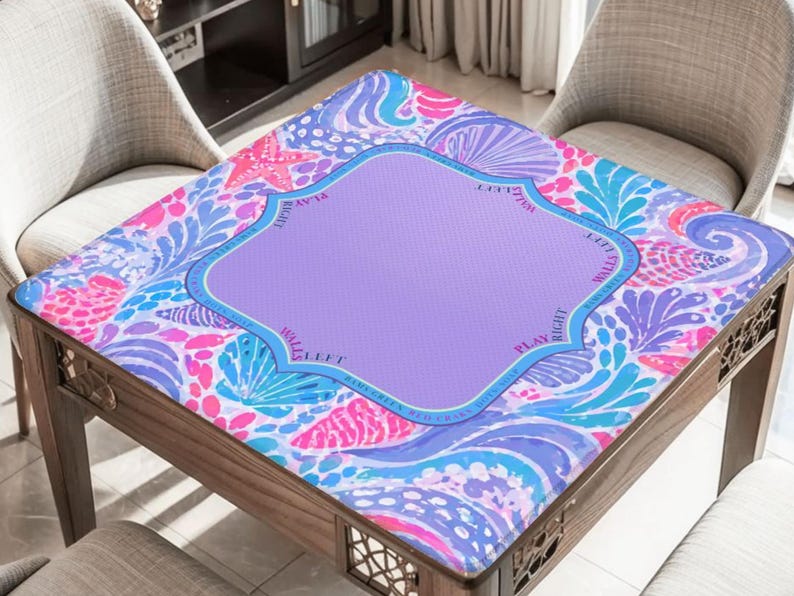 K&ouml;nnte beinhalten: Ein quadratischer Tisch mit einer bunten, gemusterten Tischplatte. Das Design zeigt eine lila Mitte mit einem dekorativen Rand, umgeben von einer lebendigen Mischung aus rosa, blauen und wei&szlig;en Blumen- und Muschelmotiven. Der Tisch ist aus Holz.