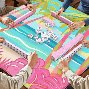 May include: A group of people playing mahjong on a table with a tropical design. The table has a colorful pattern with pink, blue, and green elements. The mahjong tiles are white with black symbols, and the racks are pink.