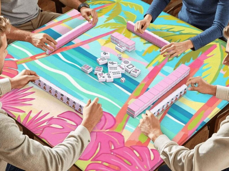 May include: A group of people playing mahjong on a table with a tropical design. The table has a colorful pattern with pink, blue, and green elements. The mahjong tiles are white with black symbols, and the racks are pink.