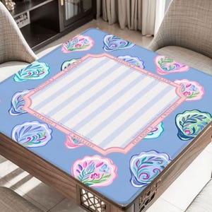Puede incluir: Una mesa cuadrada de madera con una tapa azul estampada. La tapa presenta un fondo azul claro con dise&ntilde;os de conchas marinas y un &aacute;rea central con rayas blancas y azul claro. La mesa est&aacute; rodeada por dos sillas.
