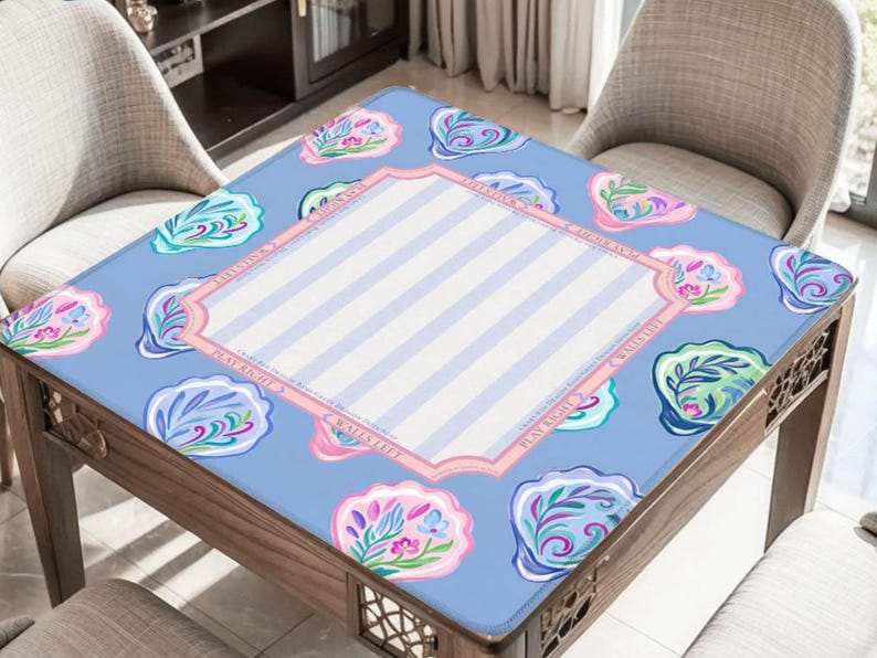 Puede incluir: Una mesa cuadrada de madera con una tapa azul estampada. La tapa presenta un fondo azul claro con dise&ntilde;os de conchas marinas y un &aacute;rea central con rayas blancas y azul claro. La mesa est&aacute; rodeada por dos sillas.