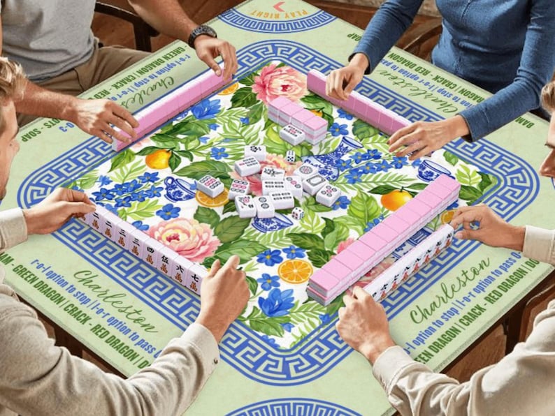 May include: A group of people playing Mahjong on a floral-patterned table. The game includes pink tile racks, white tiles, and dice. The table has a green and blue floral design with the word "Charleston" printed on it.