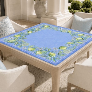 May include: A square wooden table with a light blue surface, adorned with a border of green lemons and white flowers. The table is surrounded by four cream-colored chairs with light blue pillows. The table is approximately 40 inches square.