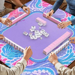 K&ouml;nnte beinhalten: Ein quadratischer Mahjong-Tisch mit einer lila Spielfl&auml;che und einem farbenfrohen floralen Rand. Rosa und wei&szlig;e Mahjong-Steine und -Racks sind auf dem Tisch angeordnet. Mehrere Personen spielen das Spiel, ihre H&auml;nde sind sichtbar.