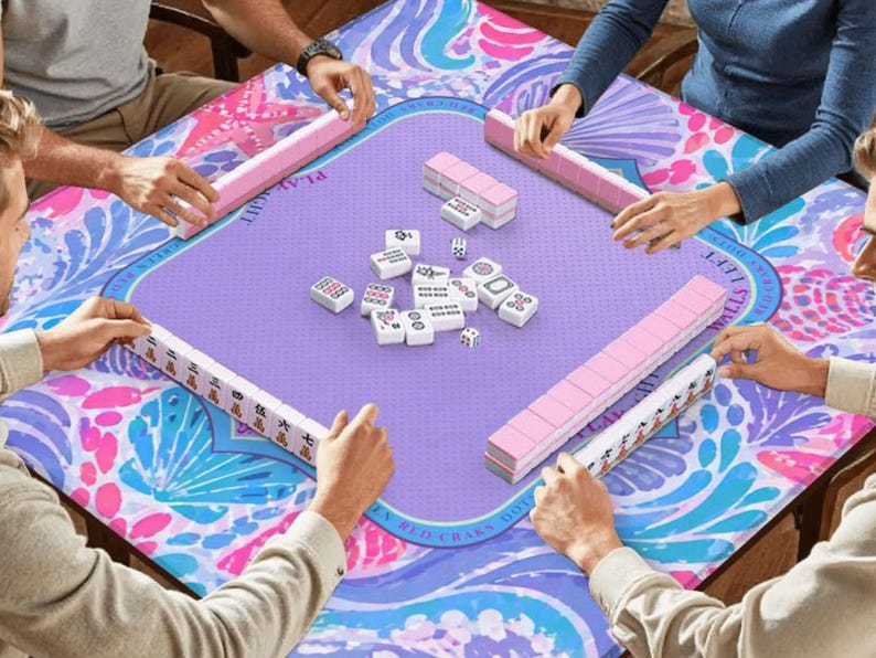 K&ouml;nnte beinhalten: Ein quadratischer Mahjong-Tisch mit einer lila Spielfl&auml;che und einem farbenfrohen floralen Rand. Rosa und wei&szlig;e Mahjong-Steine und -Racks sind auf dem Tisch angeordnet. Mehrere Personen spielen das Spiel, ihre H&auml;nde sind sichtbar.