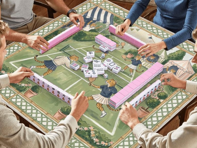 May include: A group of people playing mahjong on a decorative table. The table features a green tennis court design with cheetah illustrations. Pink and white tiles are arranged around the table. The players are holding pink and white tiles.