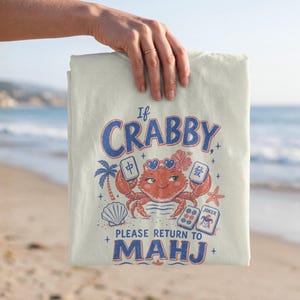 May include: A cream-coloured t-shirt featuring a graphic of a red crab wearing heart-shaped sunglasses, surrounded by mahjong tiles, a palm tree, a seashell, and a starfish. The text reads: If CRABBY, PLEASE RETURN TO MAHJ.