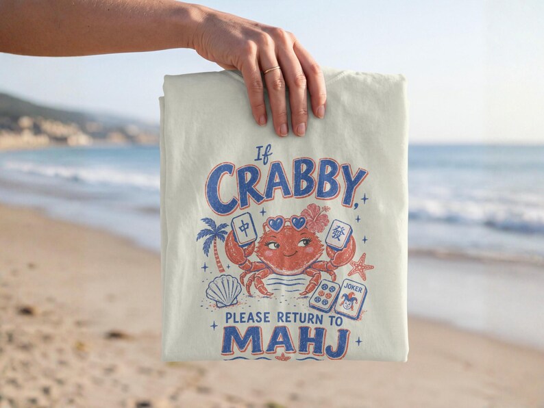 May include: A cream-coloured t-shirt featuring a graphic of a red crab wearing heart-shaped sunglasses, surrounded by mahjong tiles, a palm tree, a seashell, and a starfish. The text reads: If CRABBY, PLEASE RETURN TO MAHJ.