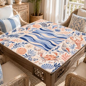 Pu&ograve; includere: Un tavolino quadrato in legno con un piano a mosaico con un design di vita marina. La superficie del tavolo presenta un motivo centrale di onde blu, circondato da illustrazioni di conchiglie, granchi e coralli nei toni del blu, arancione e bianco. Sedie in vimini con cuscini coordinati circondano il tavolo.